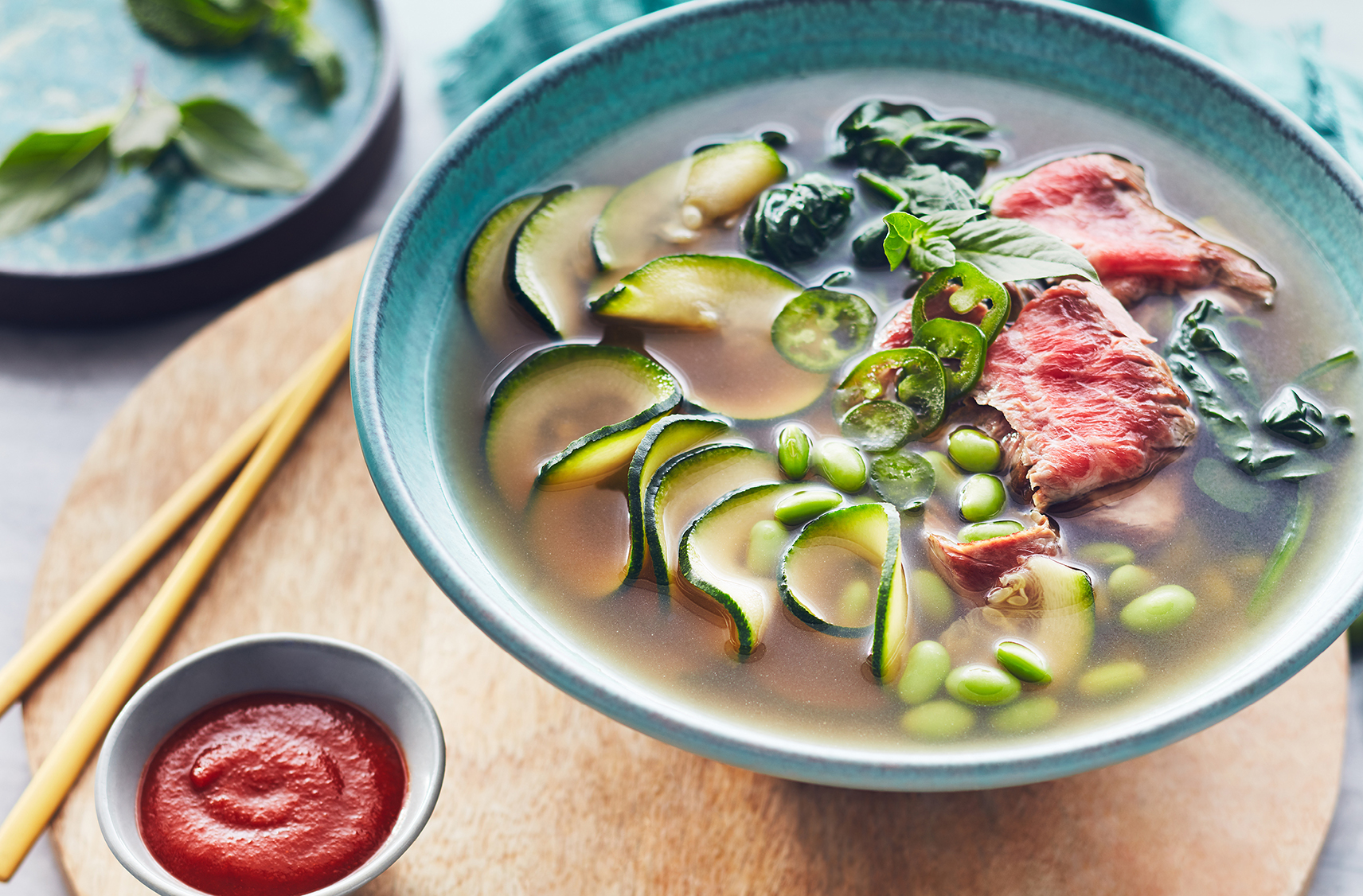 Chopsticks rest by a bowl of broth with zucchini noodles, red beef and veg
