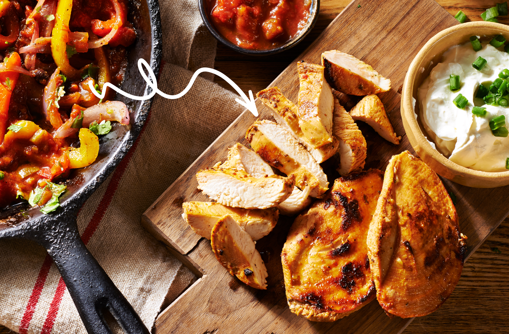 A roasted chicken sits on a cutting board with slices remove next to a cast iron fry pan with fajita ingredients