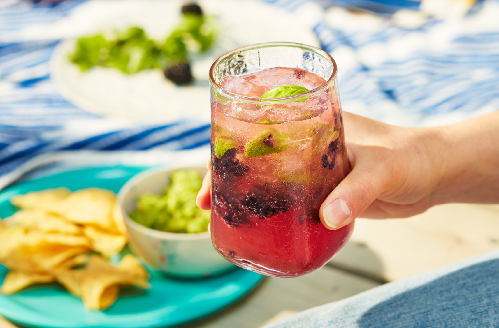 A hand holding a glass containing PC Organics Sparkling Water Kefir with blackberries and basil.  A plate of tortillas and guacamole are seen at a distance.