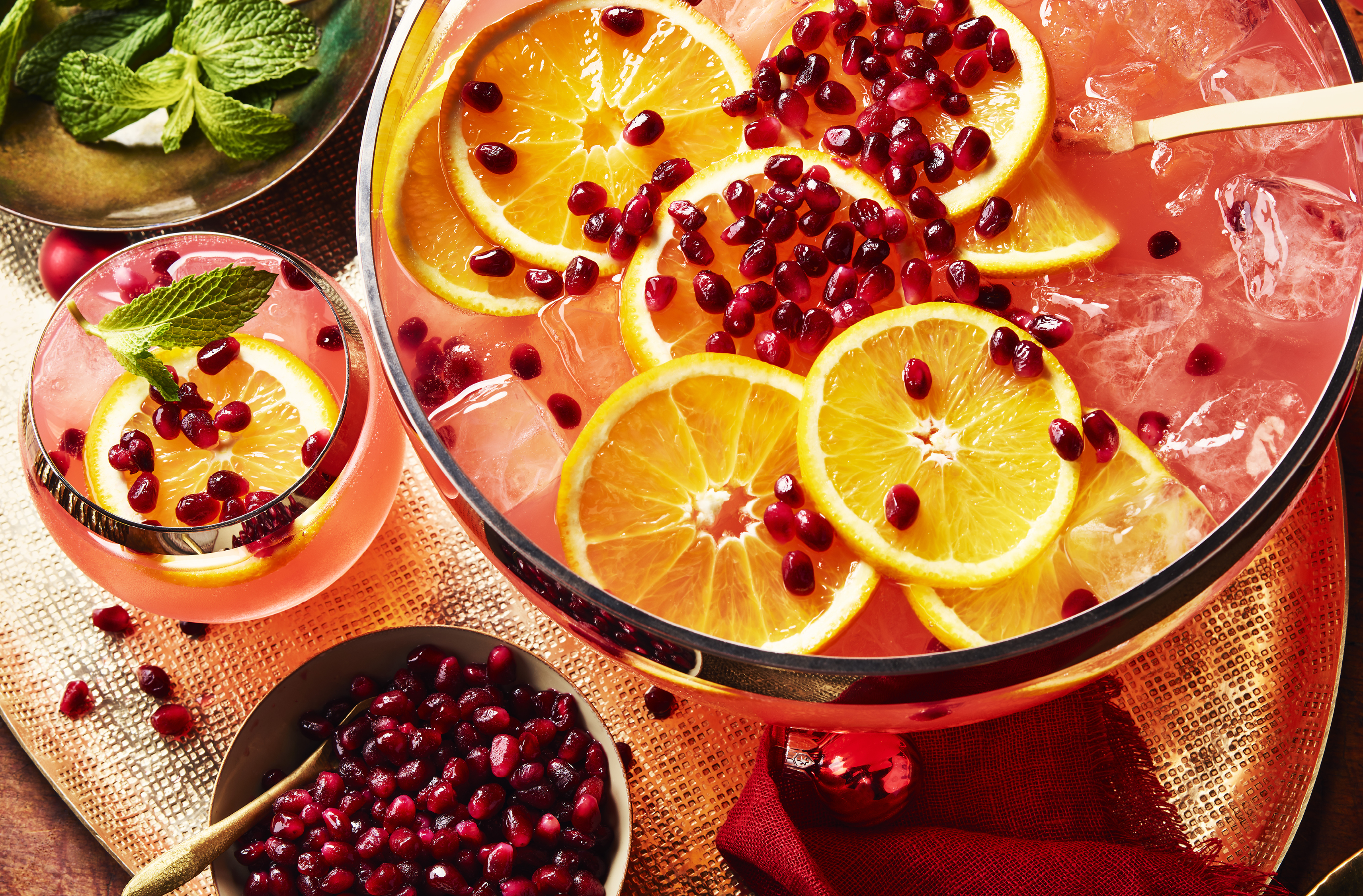 Pomegranate seeds, orange and ice float in a punch bowl beside mint leaves