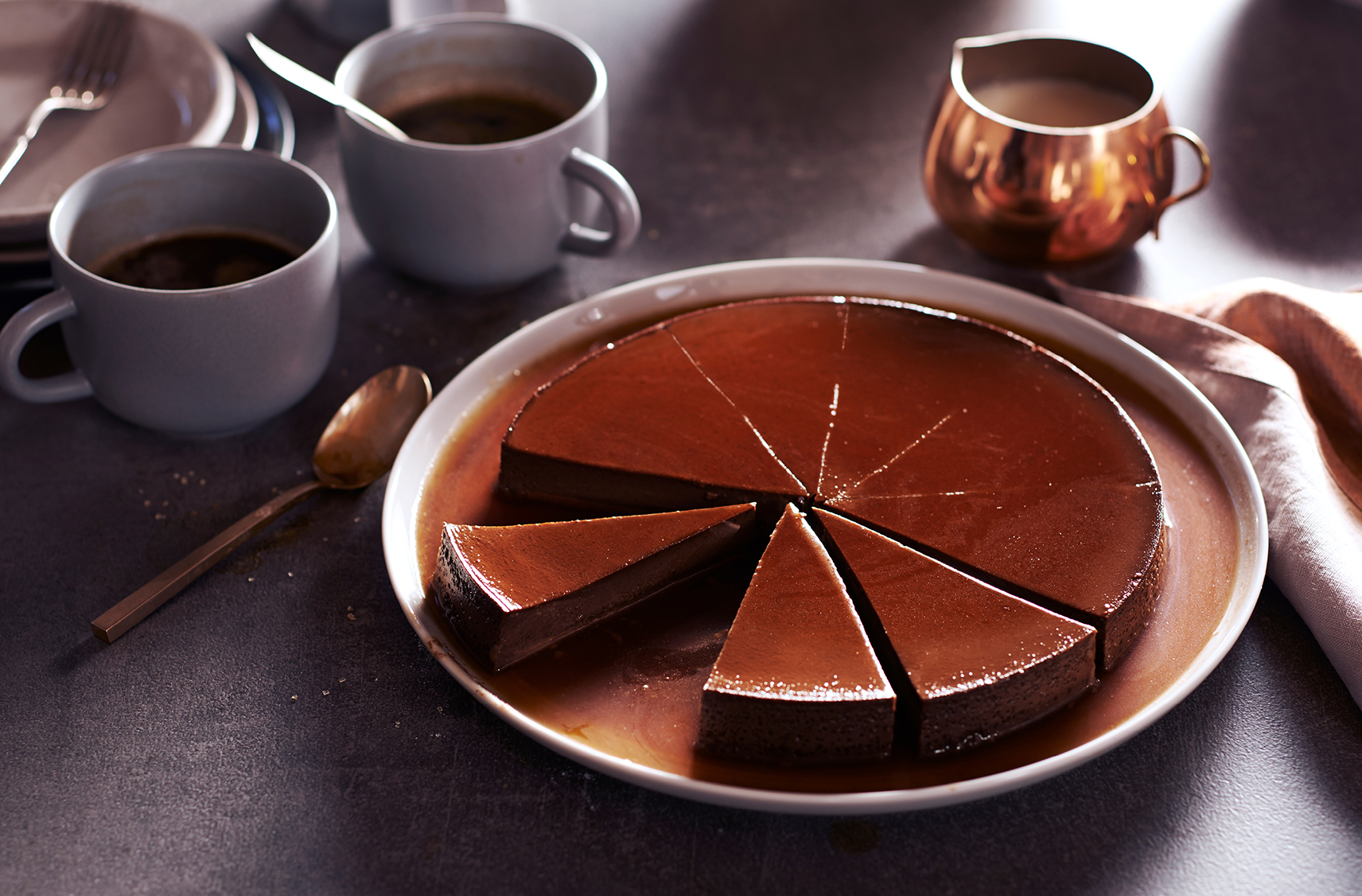 Un flan au chocolat coupé en 8 pointes dont une pointe est manquante à côté de tasses d'espresso et d'un pot de crème