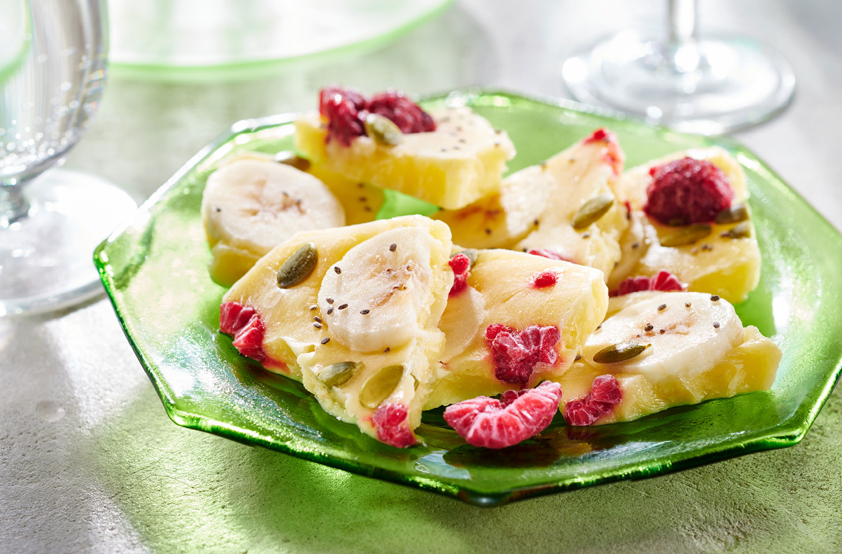 A glass plate filled with large pieces of frozen yogurt barks which contains slices of bananas, pieces of raspberries and pumpkin seeds.