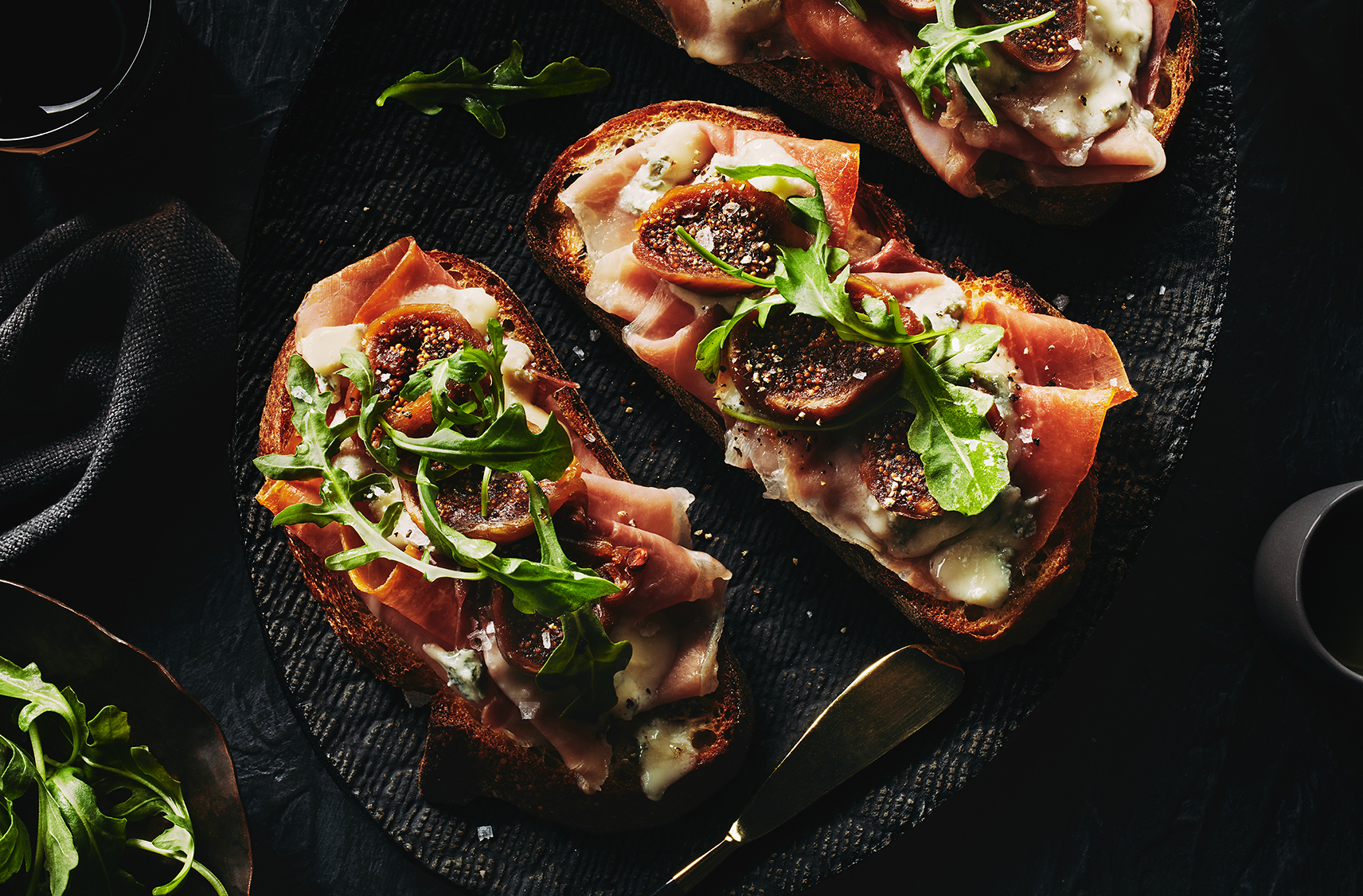 two slices of gorgonzola and prosciutto tartines placed on a black decorative plate.  Tartines are topped with some arugula

