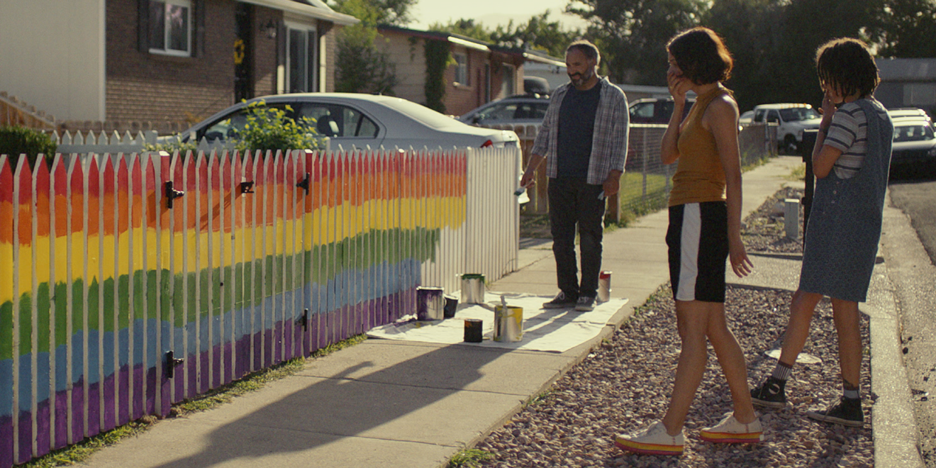 Three people stand on a sidewalk, admiring a white picket fence painted in vibrant rainbow colors. Various paint cans and supplies are scattered on the ground. The scene is set in a residential neighborhood with sunlight casting shadows on the ground.