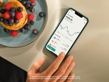 Mobile laying on a table and displaying crypto in the N26 app. Next to it, there is a pancake with raspberries and some grapes. 