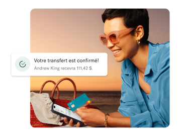 Femme sur la plage regardant son mobile et tenant sa carte N26 You. Au premier plan se trouve une notification de paiement transfrontalier.