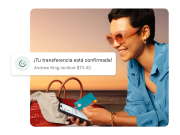 Mujer en la playa mirando su móvil y sosteniendo una tarjeta N26 You. En primer plano aparece la notificación de un pago transfronterizo.