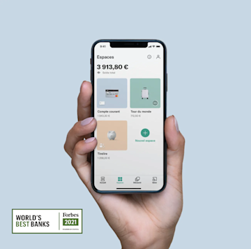 Une main tenant un iPhone X dans la main avec l'écran Spaces ouvert sur l'application N26 avec le logo Forbes de la meilleure banque.