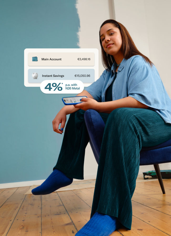 Woman dressed in blue, sitting, and holding her cell phone with the N26 application open. In the foreground, image of the balance in the main account and in the savings account.