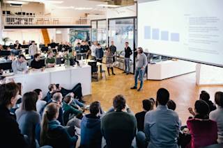 A person talking to a large group of people in front of presentation.
