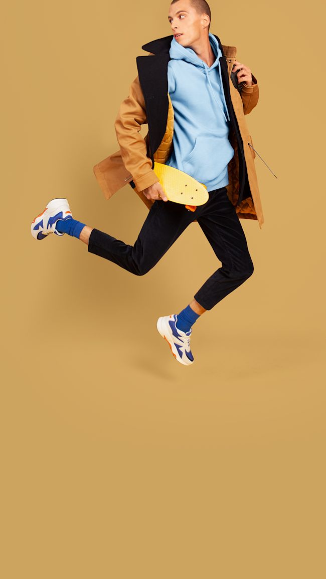 A young man dressing in a light brown jacket and a light blue hoodie holding a plastic penny board.