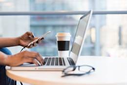 Person working on a table with a laptop, smartphone, pair of glasses and a cup of coffee on it.