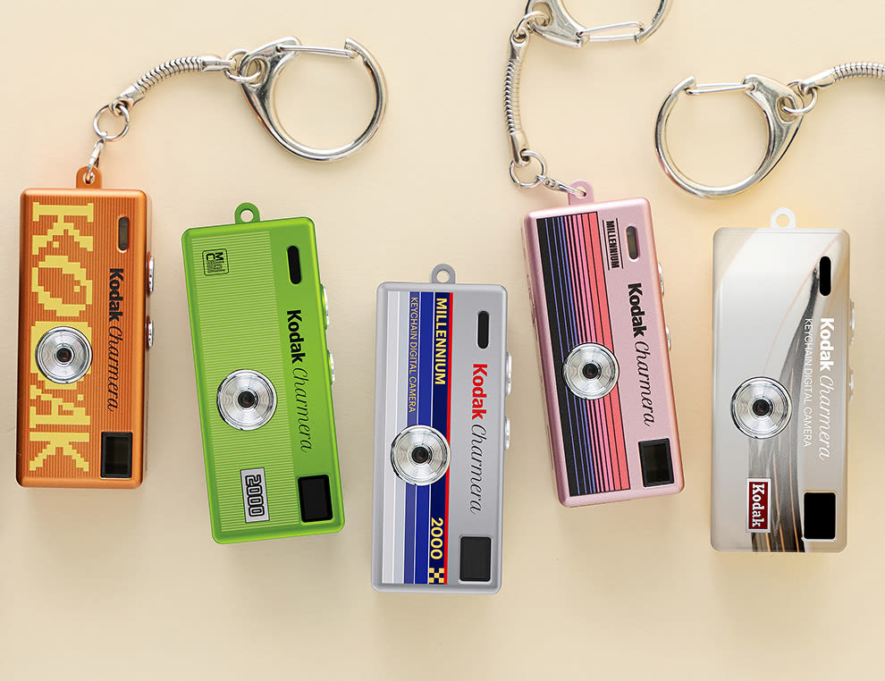 Five colorful Kodak-branded miniature camera keychains in orange, green, blue, pink, and silver on a beige background.