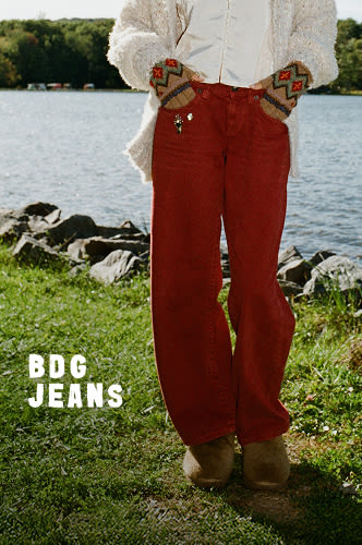 Woman in white textured cardigan and black wide-leg jeans standing by lakeside with green grass and blue sky background.