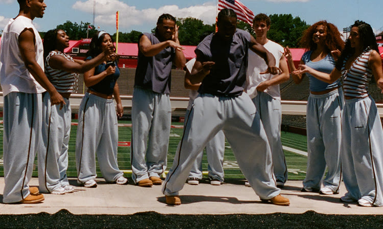 Group of diverse young people in casual athletic wear standing together on a football field with stadium seating in background.