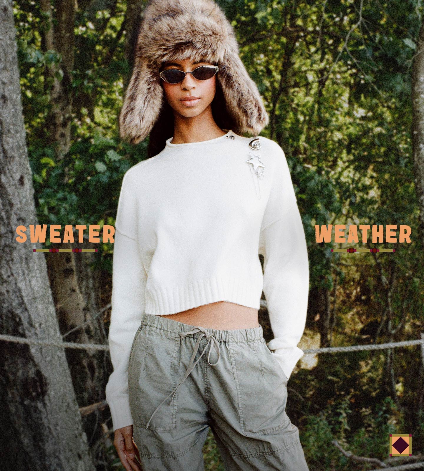 Woman wearing white cropped sweater, gray cargo pants, fur trapper hat, and sunglasses in forest with "Sweater Weather" text overlay.
