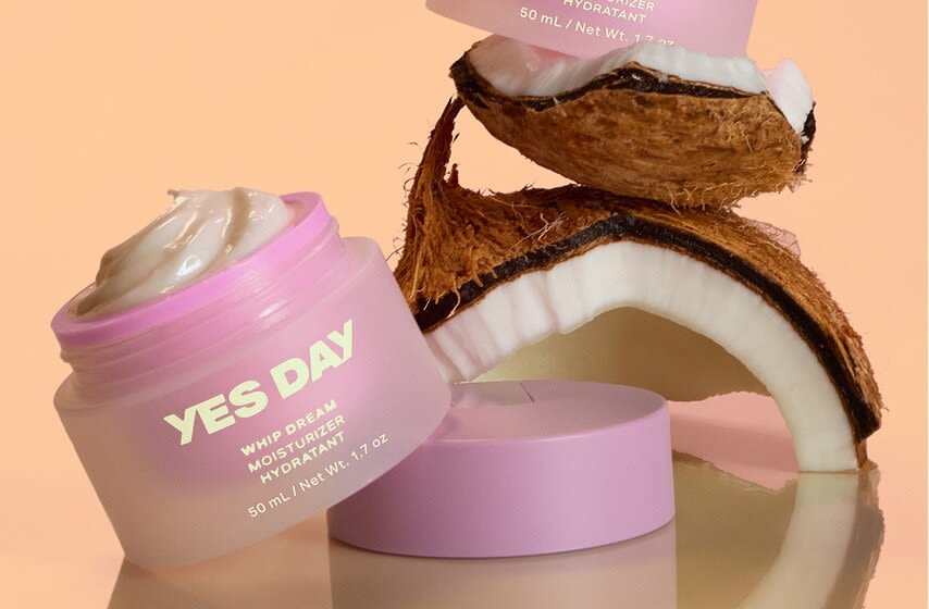 Pink YES DAY moisturizer jars with whipped cream texture displayed alongside cracked coconut halves on peach background