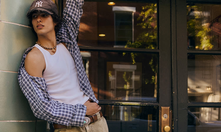 Two men modeling casual outfits: one in checkered shirt and cap by tiled wall, another in red striped sweater outdoors.