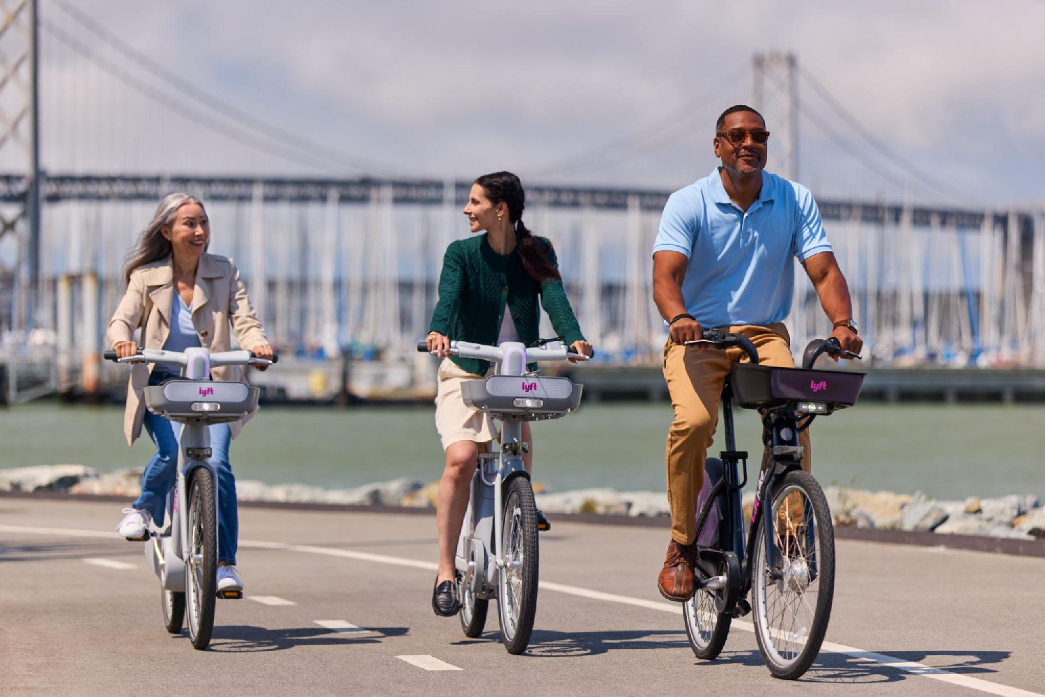 Three people ride Lyft Bike bikes along a waterfront path with a bridge in the background.