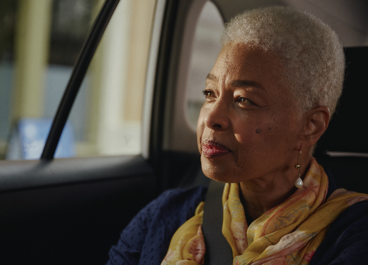 Older woman with short gray hair in a car, wearing a yellow scarf, looking out window..