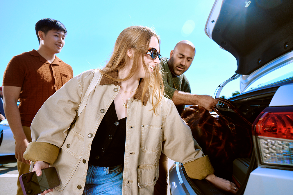 Friends loading luggage into an open car trunk in a sunny parking lot.