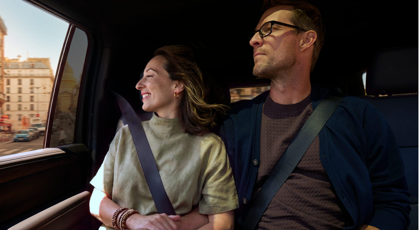 Smiling couple wearing seatbelts in a car, looking out window at city buildings.