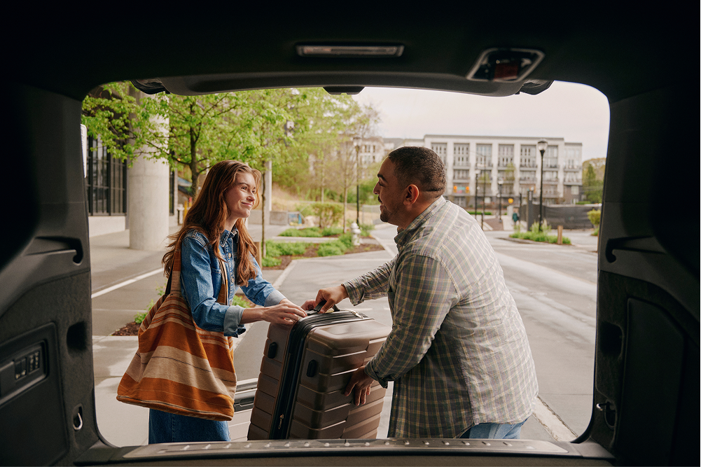 Man helps woman load a suitcase into an SUV trunk on a city street.