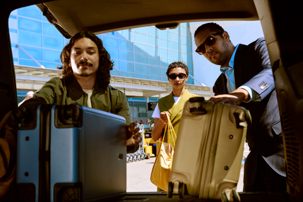 Three travelers load blue and beige suitcases into a car trunk outside an airport.