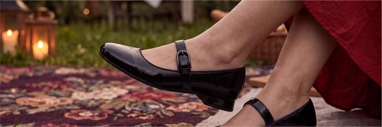 Nahaufnahme der Beine einer Person in einem roten Kleid, die auf einem gemusterten Teppich im Garten sitzt und schwarze, glänzende Mary-Jane-Ballerinas mit Riemchen trägt; im Hintergrund leuchten Laternen.