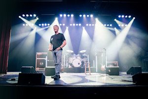 A man stands on a stage with instruments behind him and spotlights on around him.