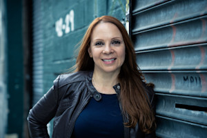 Close-up of a woman with long hair and wearing a leather jacket leaning against a building.