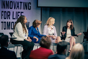4 women sit in chairs on a stage and one is speaking while the others look toward her.