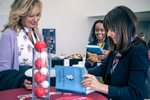 Woman signs a book at a table for another woman.