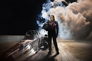 Woman stands next to a race car holding a helmet and wearing race gear.