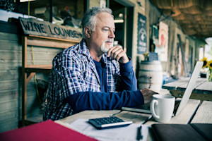 Older man sits at a table looking at a laptop with a calculator, paperwork, and a coffee mug.