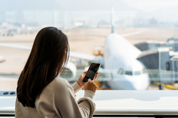 A woman holding a mobile phone in her hand looking out of a terminal window viewing a plane parked at a gate.