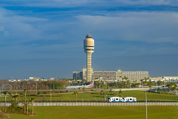 Outside, looking at Terminal AB with control tower in the background. The sky is partly cloudy with blue sky. Upfront is a Terminal Link passing by. In the mid ground is an airline plane taxiing in front of the control tower.