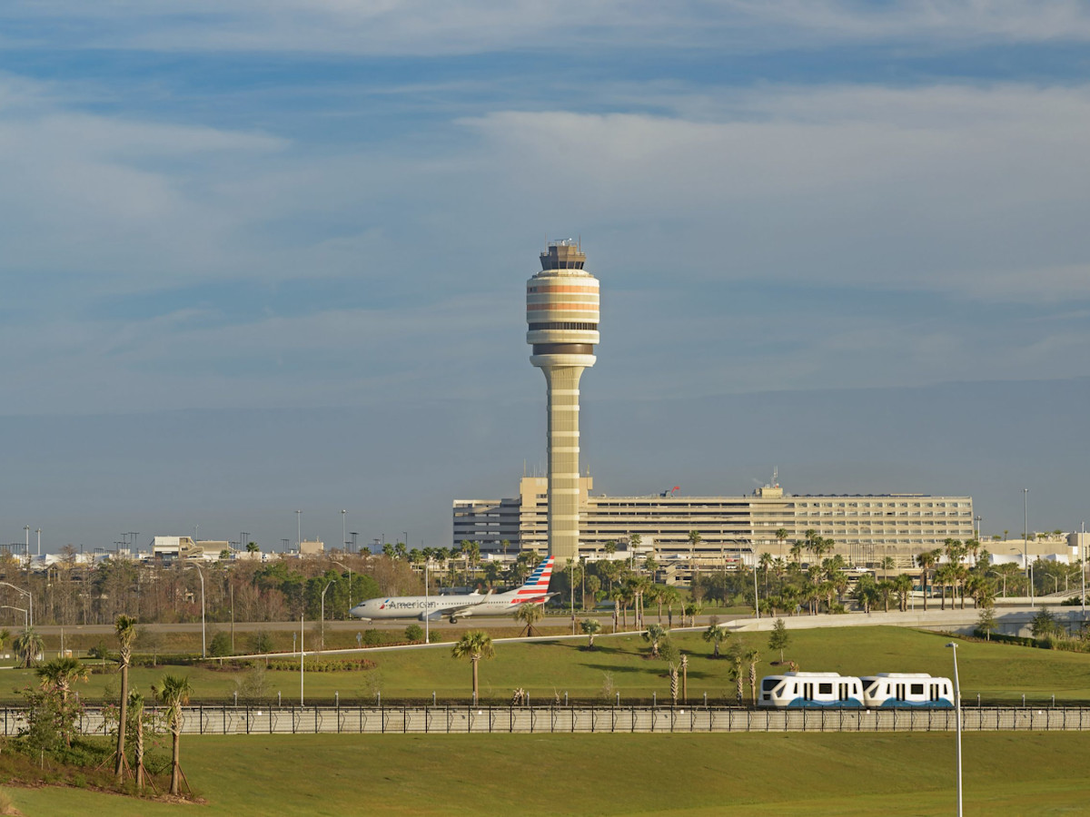 Outside view of the MCO Airport Terminal AB Control Tower. In the middle ground is a plane taxiing across a bridge, In the foreground is a Gate link moving along the track.