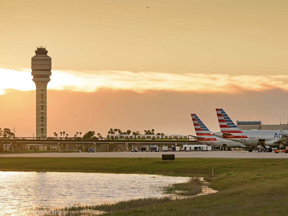 A view of the MCO tower in the background with a sunrise. Two airplanes parked at a Terminal AB gate in the foreground.