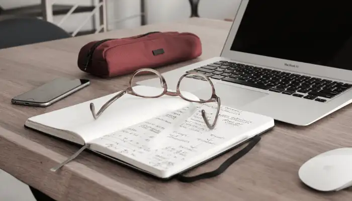 a desk with a laptop, book, mouse, glasses, phone and pencil case