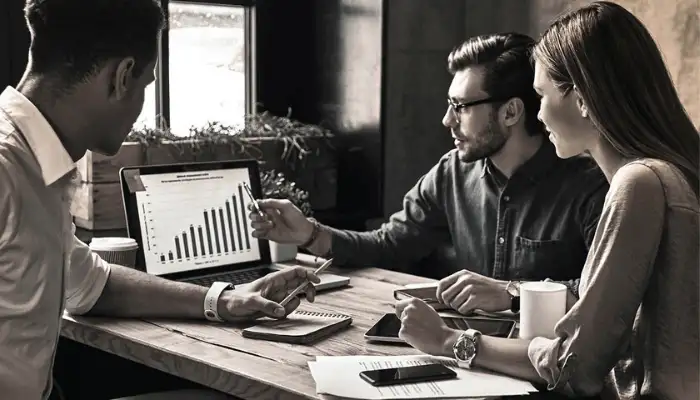 three people sitting around a table having a meeting and looking at a laptop