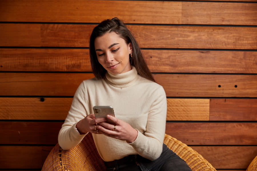 Image of a young woman in her early 20s sitting outside. She's looking down at her phone and smiling.