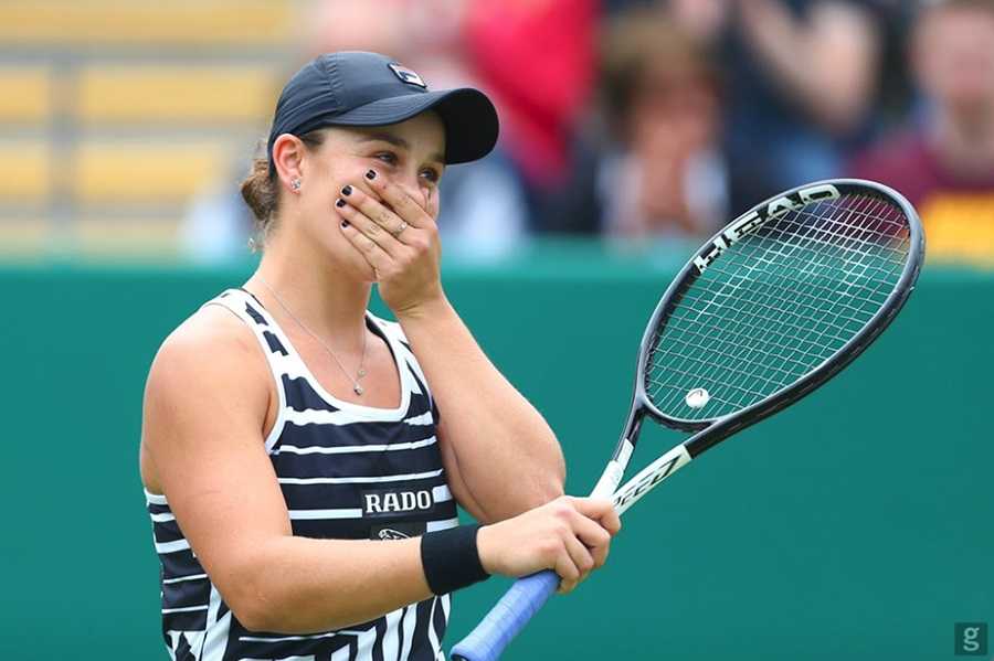 Ash Barty smiling with hand covering mouth and holding a tennis racquet