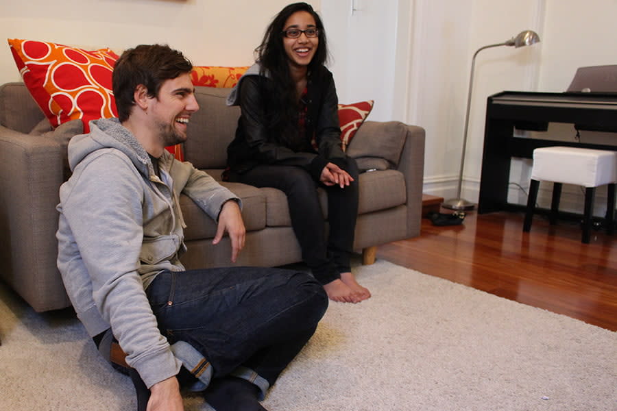 Image of a young man and woman relaxing in the living room at home. The guy is sitting on the floor with his arm on the couch, next to where the woman is sitting. They are both laughing.