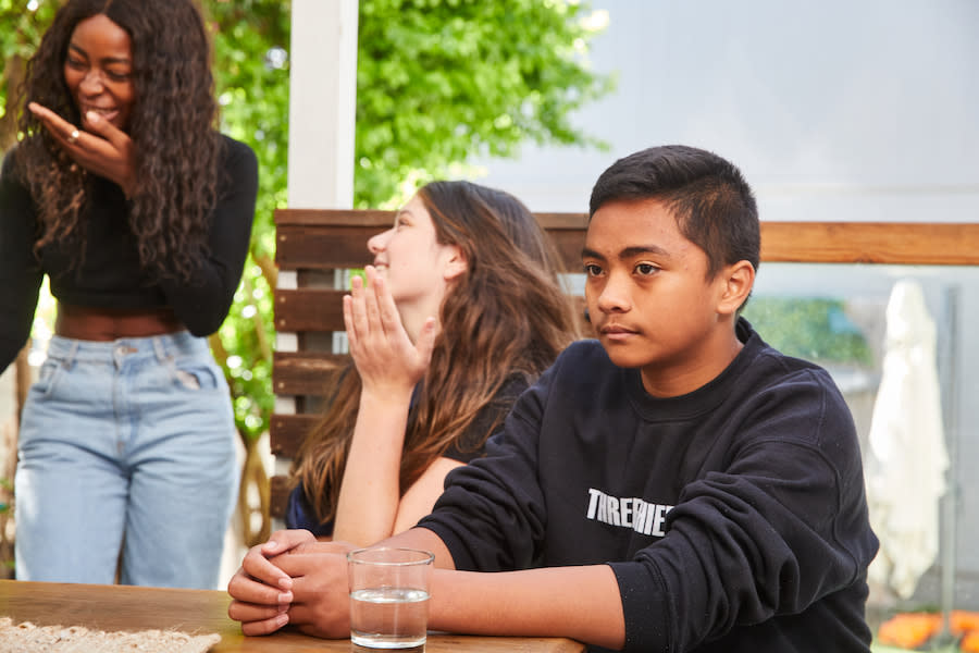 Image of a teenage boy and two teen girls. The boy is sitting at a table looking into the distance, he has a sad look on his face. The teen girls are standing behind the boy chatting to each other and laughing.