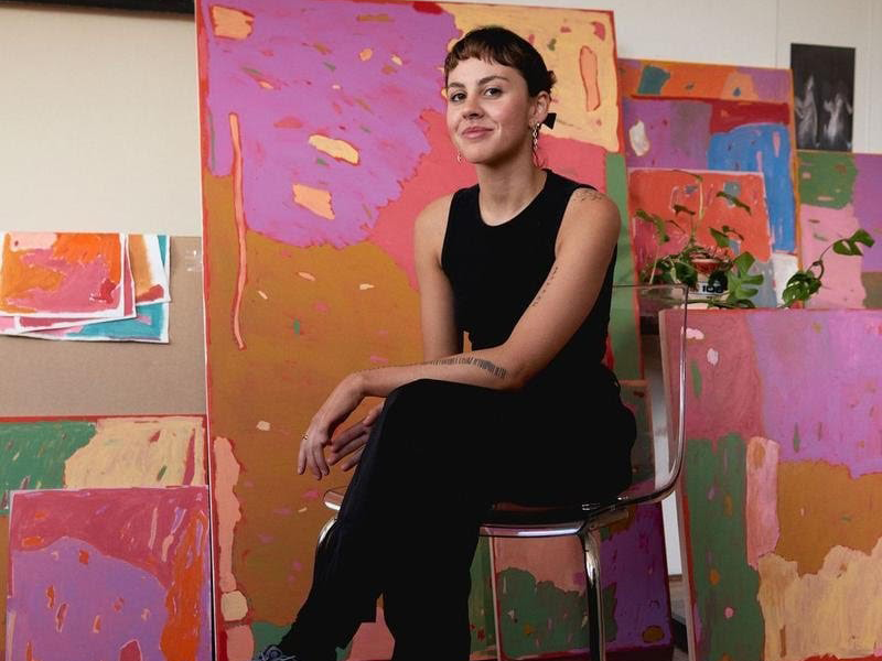 Image of Tiarna, a smiling woman with light skin and dark brown hair. She is wearing all black and sitting in a chair in front of a number of colourful paintings that are leaning against a wall.