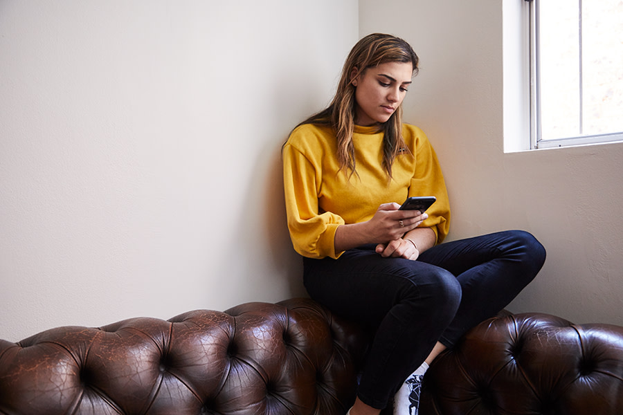 Image of a girl sitting on a lounge, looking at her phone. She has a concerned look on her face.