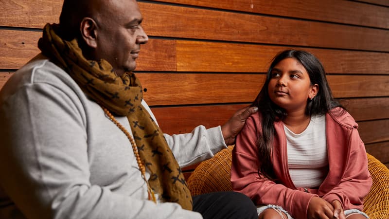 Image of an adult man and his younger teenage daughter sitting together. The man has black skin and is resting his hand on his daughter's shoulder. The young girl has brown skin and long black hair. She is looking up at her dad attentively.