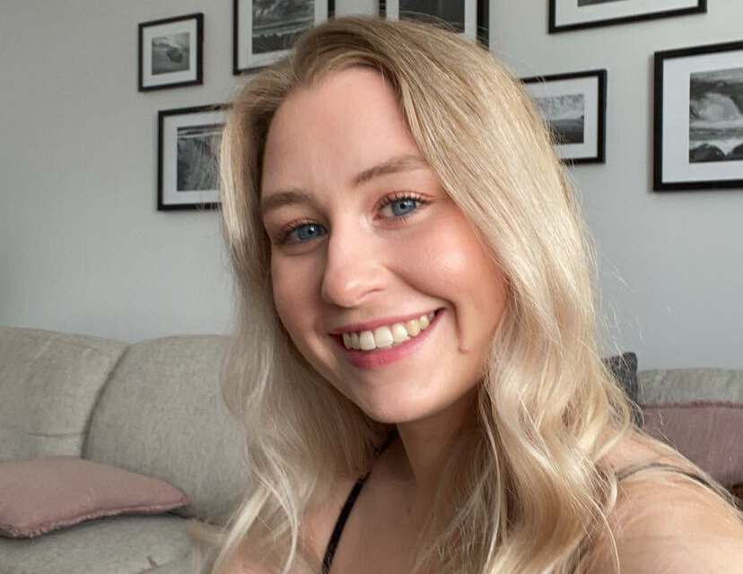 Selfie image of Annie sitting in a lounge room. She has light skin, blue eyes, and long blonde hair. She is smiling widely at the camera.