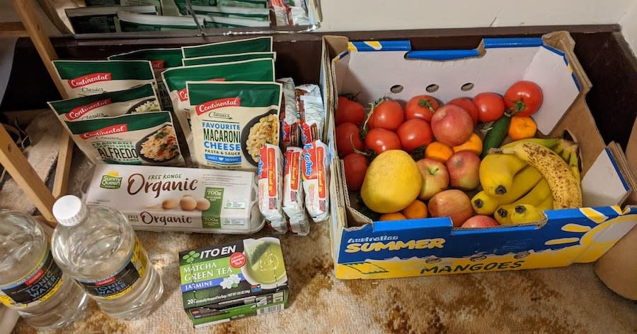 Image of a care package of food. In the package there is pasta packets, noodles, eggs, fruit, vegetables, and tea.